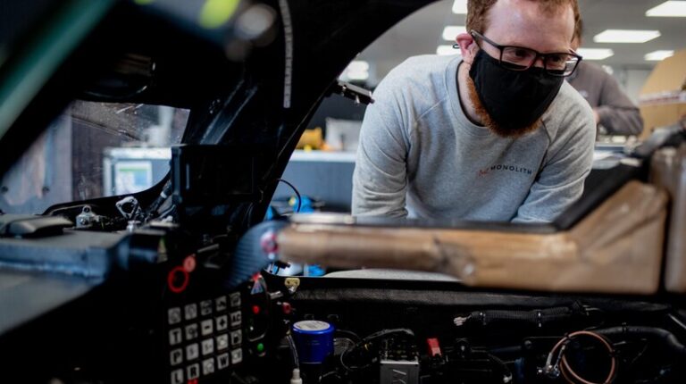 An automotive engineer wearing a face mask looks through the open window of a vehicle. The dash contains various testing equipment.