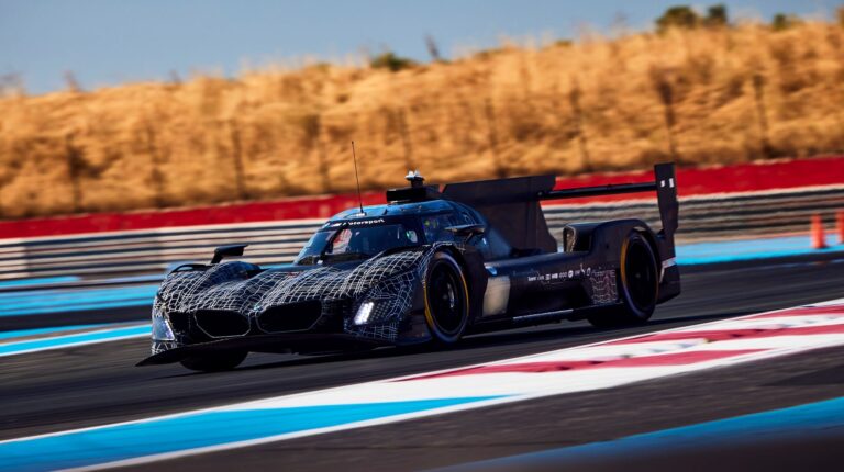 The BMW M Hybrid V8 takes a turn at high speed on a racetrack with hay bales in the background