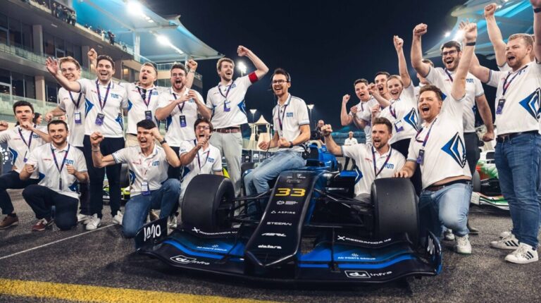TUM team members celebrate their win at the second A2RL Grand Final with the car parked between them and the grandstand rising up on either side into the night sky