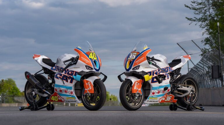 Two BMW Motorrad Motorsport bikes parked on an empty racing track, with orange, white and blue OrangeCat Racing livery