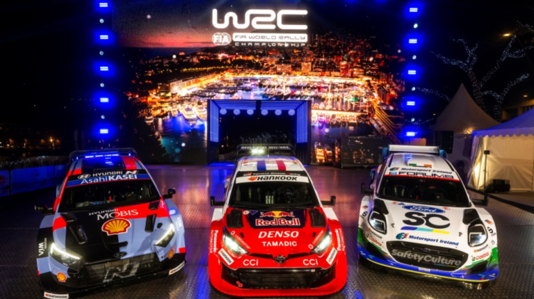 Three rally cars sit on a promotional stage in front of WRC logo and branding at the season opener in Monte Carlo. The cars are red, black and white.