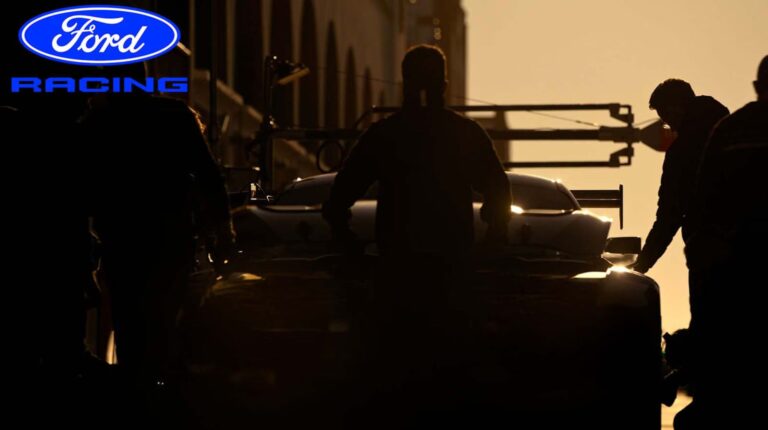 A silhouette of a hypercar on the track against a setting sun. Engineers stand at the side and Ford logo is in the top left hand corner.