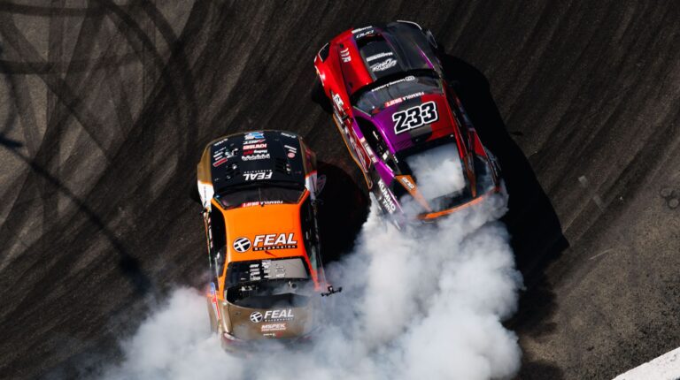 Two Formula Drift cars - one red, one orange and black - in competition, with white smoke coming out the back of the vehicles.